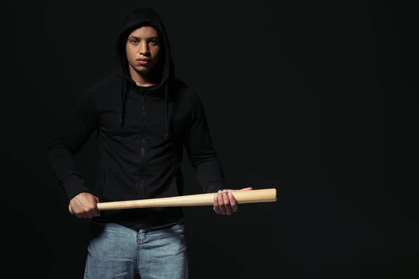 African american man in hoodie holding baseball bat and looking at camera isolated on black - foto de stock