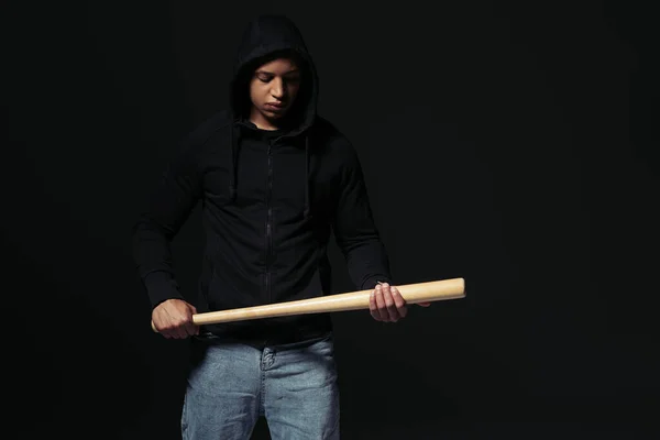 African american hooligan in hoodie looking at baseball bat isolated on black — Stock Photo