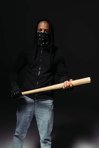 African american hooligan with covered face holding baseball bat on black background - foto de stock