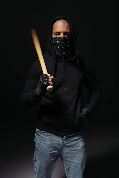 African american hooligan with scarf on face holding baseball bat on black background — Stockfoto