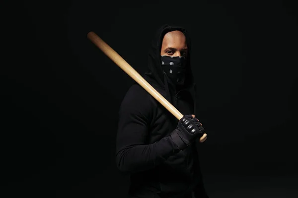 African american hooligan with mask on face holding wooden baseball bat isolated on black — Fotografia de Stock