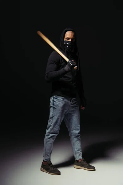 Full length of afrcan american hooligan in mask on face holding baseball bat on black background — Stock Photo