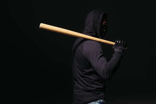 Side view of african american hooligan in mask and glove holding baseball bat isolated on black — Stockfoto