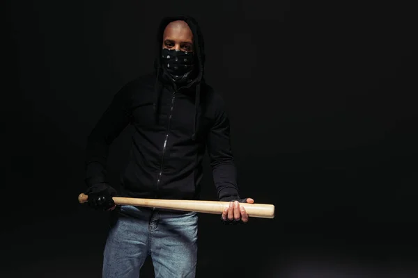 African american hooligan in gloves and mask on face holding baseball bat isolated on black — Fotografia de Stock