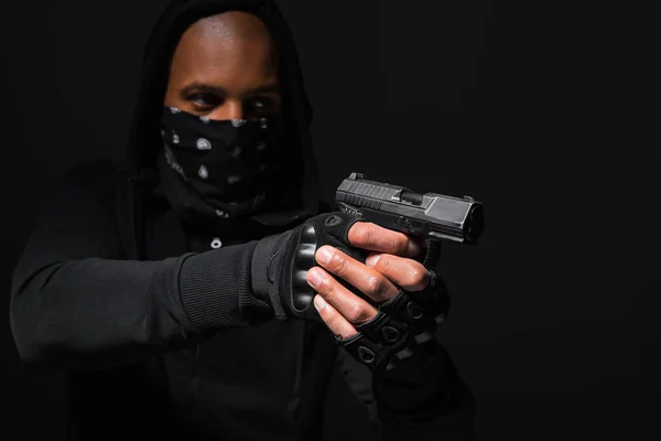 Blurred african american hooligan in mask holding gun isolated on black — Foto stock