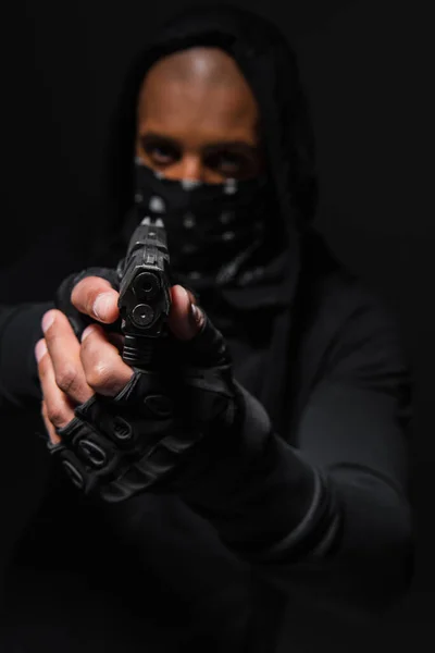 Blurred african american bandit with mask on face holding gun isolated on black - foto de stock