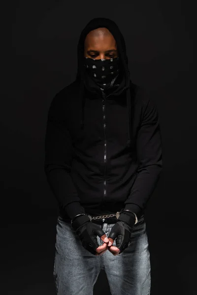 African american bandit with mask on face and handcuffs standing isolated on black - foto de stock