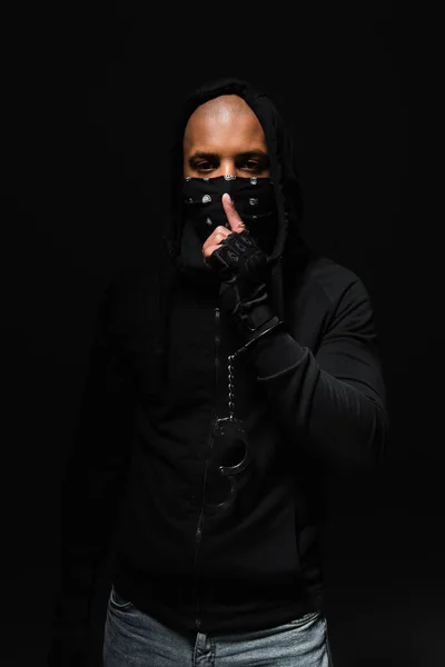 African american bandist in mask on face and handcuffs showing secret gesture isolated on black — Stockfoto