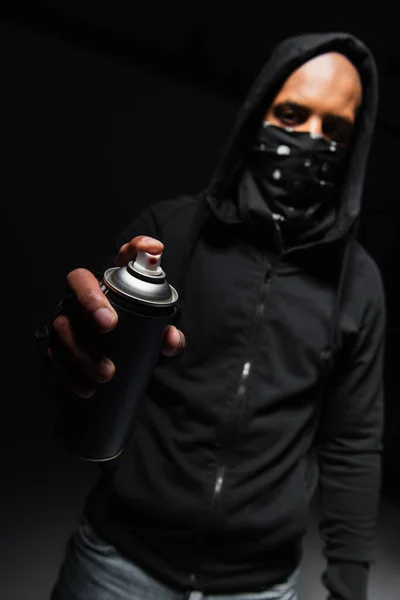 Hooligan afroamericano sfocato con maschera sul viso che tiene vernice spray su sfondo nero — Foto stock