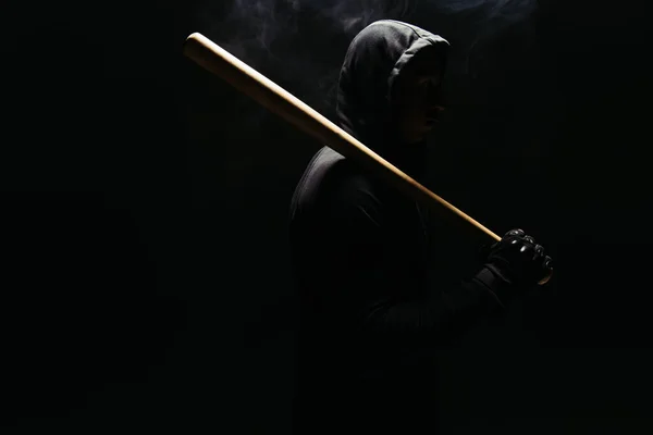 Silhouette of bandit in hoodie holding baseball bat on black background with lighting and smoke - foto de stock