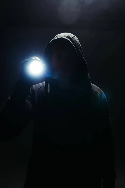 Silhouette of bandit in hoodie holding flashlight on black background with smoke — Stock Photo