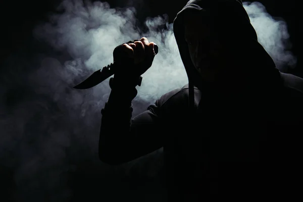 Silueta de bandido afroamericano en capucha sosteniendo cuchillo sobre fondo negro con humo - foto de stock