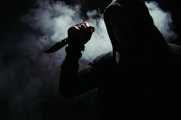 Silhouette of african american bandit in hood holding knife on black background with smoke — Stock Photo