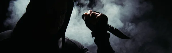 Silhouette of man in hoodie holding knife on black background with smoke, banner — Stock Photo