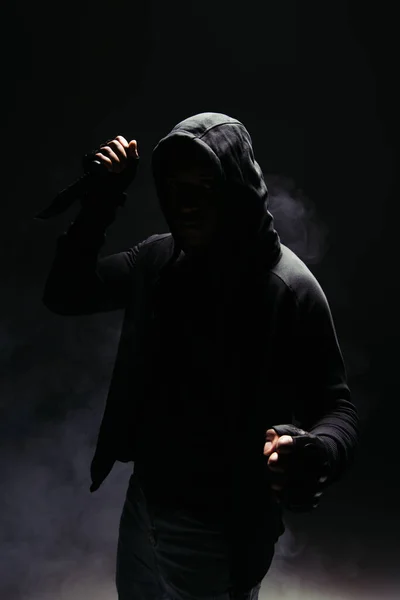 Silhouette of african american man in hood and gloves holding knife on black background with smoke — Photo de stock