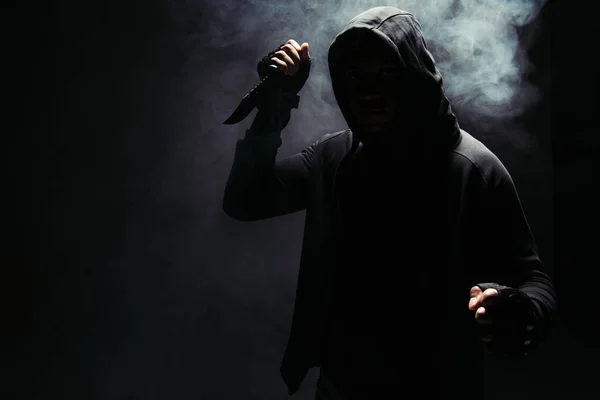 Silhouette of african american bandit in hoodie holding knife on black background with smoke - foto de stock