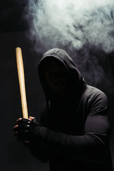 Silhouette of african american bandit in hood holding baseball bat on black background with smoke - foto de stock