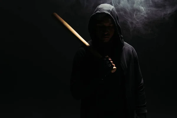 Silhouette of african american hooligan in hood holding baseball bat on black background with smoke — Stockfoto