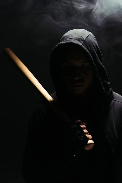 Silhouette of african american bandit in hood and glove holding baseball bat on black background with lighting and smoke - foto de stock