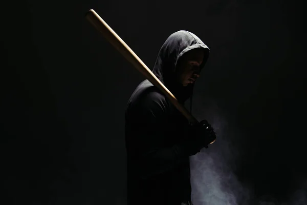 Side view of african american bandit in hood holding wooden baseball bat on black background with smoke - foto de stock