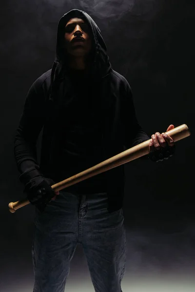 African american bandit in hood holding baseball bat on black background with smoke — Stockfoto