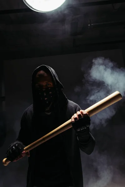 Silhouette of african american vandal with mask on face holding baseball bat on black background with smoke — Fotografia de Stock