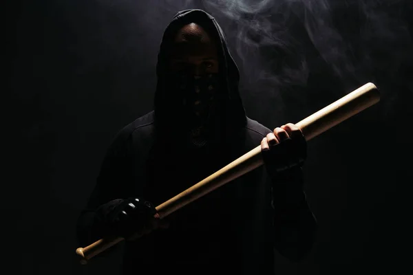 Silhouette of african american man in hoodie and gloves holding baseball bat on black background with smoke — Photo de stock