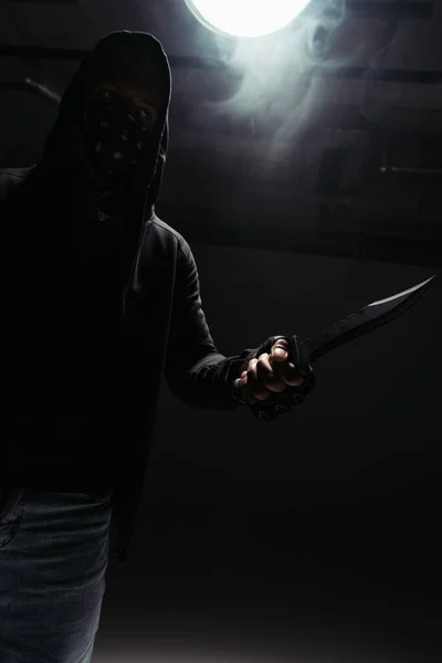 Silueta de hooligan afroamericano con oscura cara sosteniendo cuchillo sobre fondo negro con humo - foto de stock