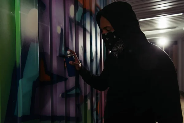 Side view of african american hooligan in hoodie and mask drawing graffiti on wall — Foto stock