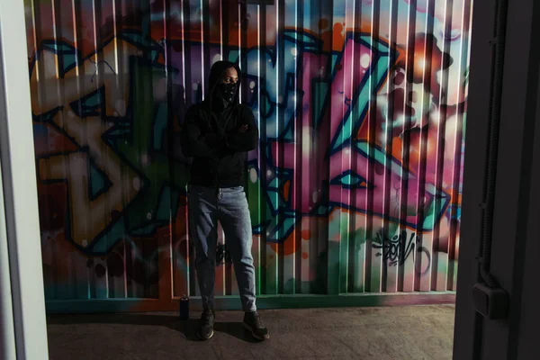 Vándalo afroamericano con máscara en la cara de pie cerca de graffiti en la pared - foto de stock