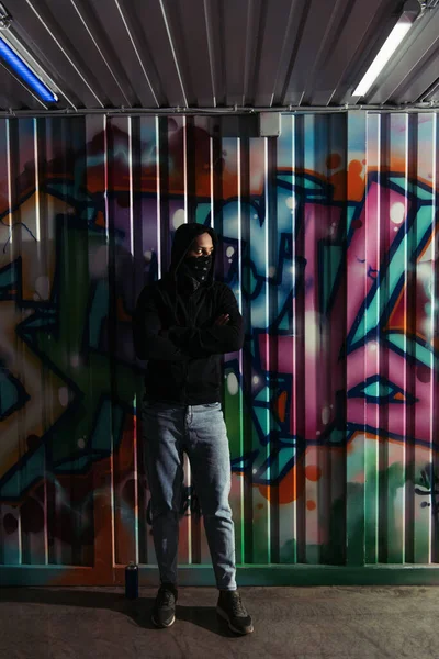 African american vandal in hoodie standing near spray paint and graffiti on wall - foto de stock