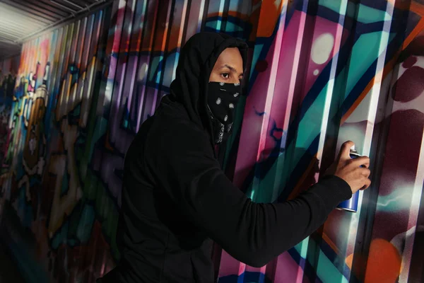 Vándalo afroamericano con bufanda en la cara dibujo graffiti en la pared - foto de stock