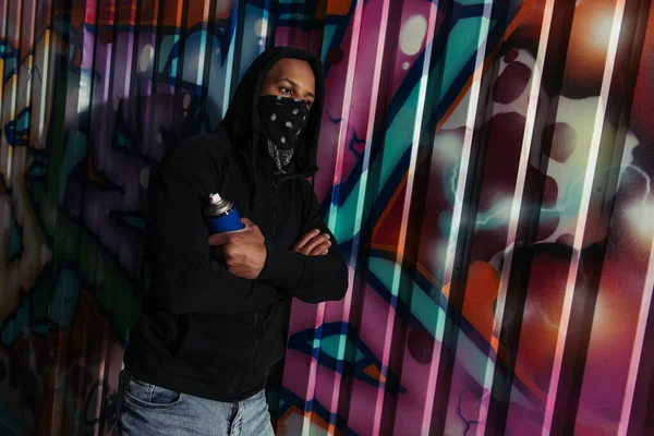 African american hooligan in hoodie holding spray paint near graffiti on wall on urban street — Foto stock