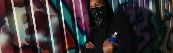 Vándalo afroamericano con cara cubierta sosteniendo pintura en aerosol cerca del graffiti en la calle urbana, pancarta - foto de stock