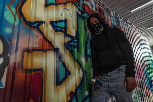 Vista de bajo ángulo del vándalo afroamericano con máscara en la cara sosteniendo pintura en aerosol cerca del graffiti - foto de stock