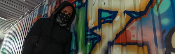 Low angle view of african american vandal looking at camera near graffiti on wall, banner — Stock Photo