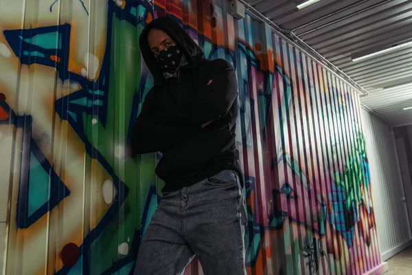 African american vandal in mask and hoodie crossing arms near graffiti on wall — Stock Photo