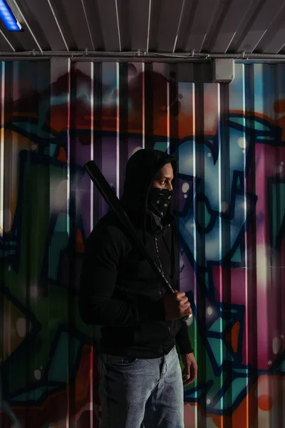 Side view of african american hooligan in mask and hoodie holding baseball bat near graffiti on wall - foto de stock