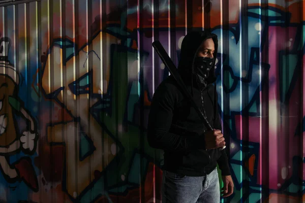 African american vandal with mask on face holding baseball bat near graffiti on urban street — Fotografia de Stock