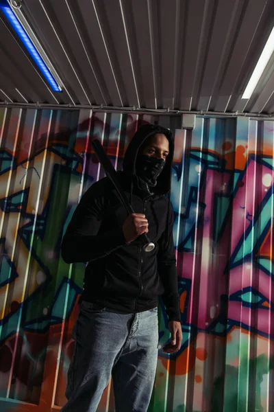 Vándalo afroamericano con máscara sosteniendo bate de béisbol cerca de graffiti en la pared - foto de stock