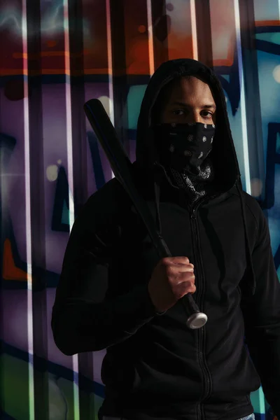 African american hooligan holding baseball bat and looking at camera near graffiti on urban street — Foto stock