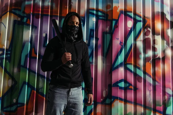 African american hooligan holding baseball bat and looking at camera near graffiti outdoors - foto de stock
