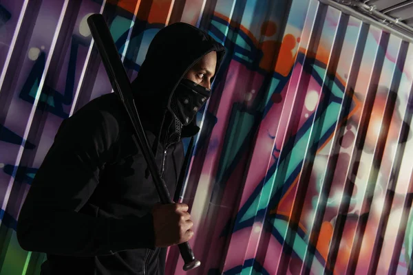 Side view of african american bandit in mask holding baseball bat near graffiti on wall — Stock Photo