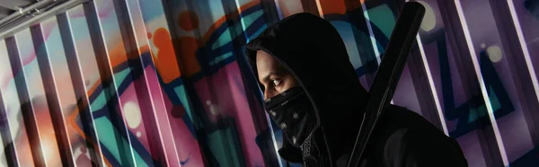Vista lateral del bandido afroamericano con máscara sosteniendo bate de béisbol cerca de graffiti en la pared, pancarta - foto de stock