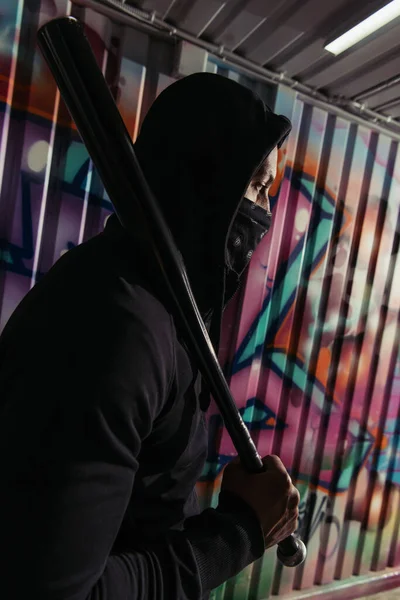 Side view of african american vandal in mask holding baseball bat near graffiti on wall — Stock Photo