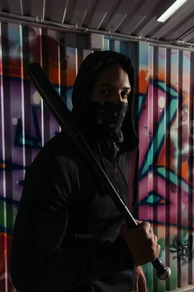 African american hooligan with baseball bat looking at camera near graffiti on wall — Fotografia de Stock