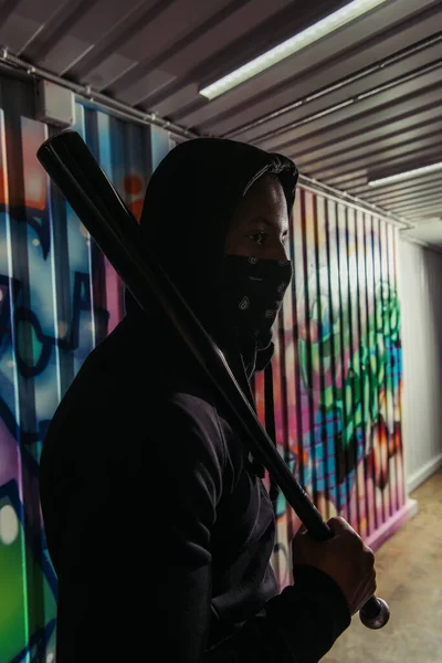 African american hooligan in mask holding baseball bat near graffiti — Stock Photo