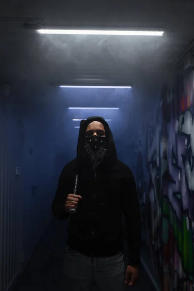 African american hooligan in mask holding baseball bat and looking at camera near lighting and graffiti — Fotografia de Stock