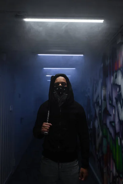 African american hooligan in mask holding baseball bat and looking at camera near lighting and graffiti - foto de stock