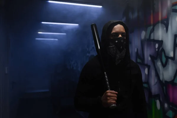 African american hooligan holding baseball bat near graffiti and smoke in garage — Stock Photo